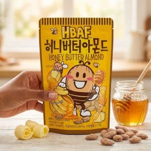 Archives Bright yellow package labeled 'Honey Butter Almond' with a smiling cartoon almond/bee character, held by a hand, on a wooden table with honey jar nearby.