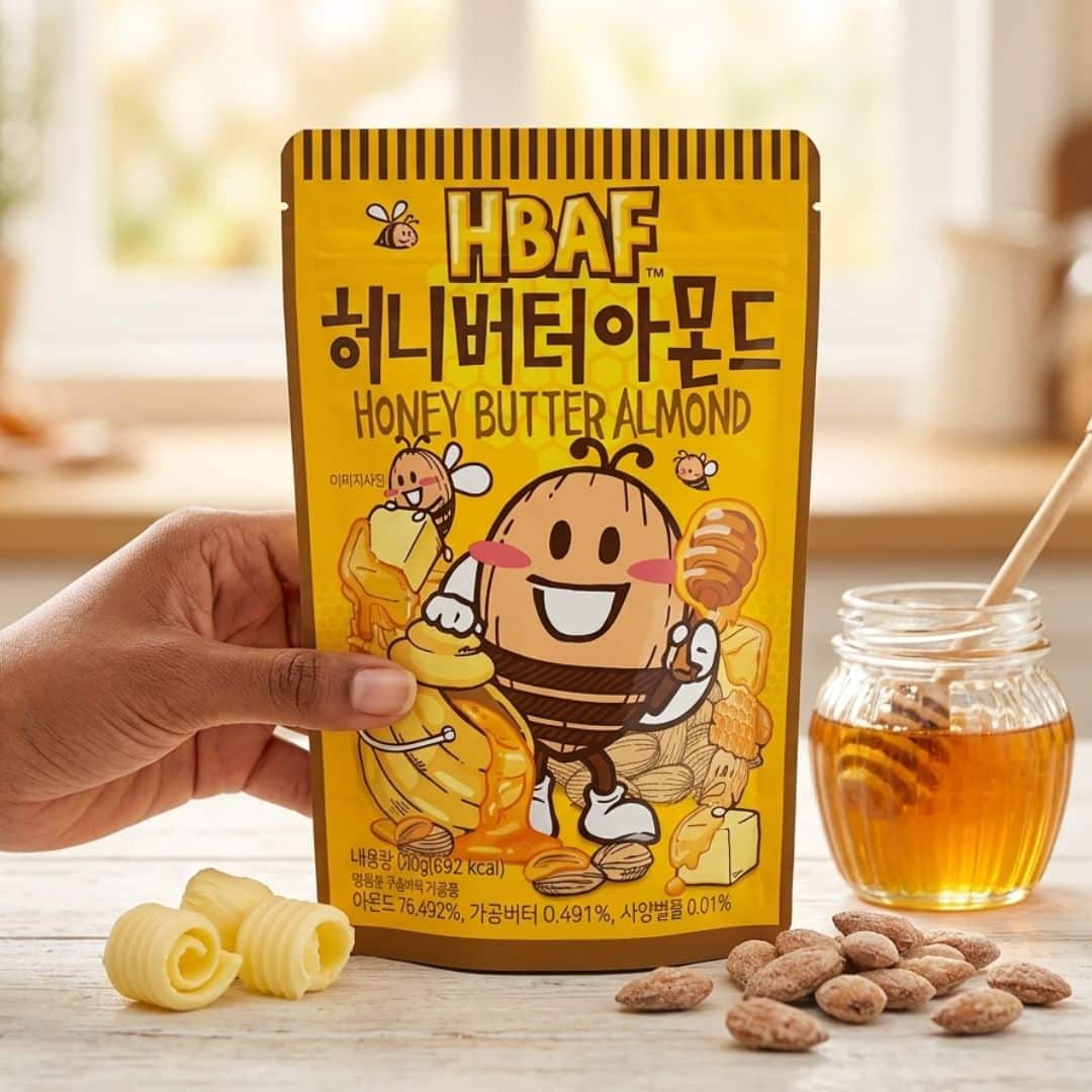 hbaf honey butter almond | Snack Affair Archives Bright yellow package labeled 'Honey Butter Almond' with a smiling cartoon almond/bee character, held by a hand, on a wooden table with honey jar nearby.