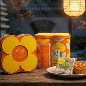 Bright orange-yellow mooncake gift tin and matching flower-shaped box labeled CASA HANA with a floral pattern, set on a wooden table beside a plate of mooncakes and a hanging lantern in warm evening light.