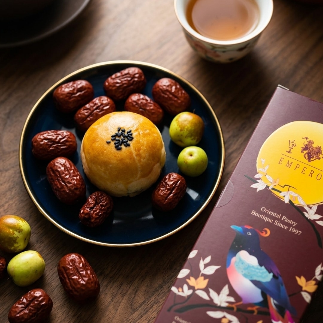 Emperor Jujube mooncake 2 | Snack Affair Archives Round pastry with black sesame seeds on a blue plate, surrounded by red dates and green plums, with a cup of tea and a decorative pastry box nearby.