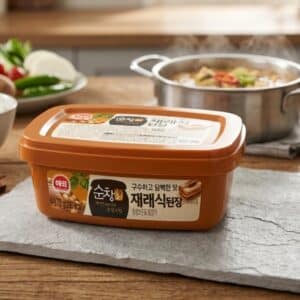 Archives Orange rectangular container of Korean miso-like paste on a kitchen table with a pot of stew in the background, label featuring Korean text 재래식된장.