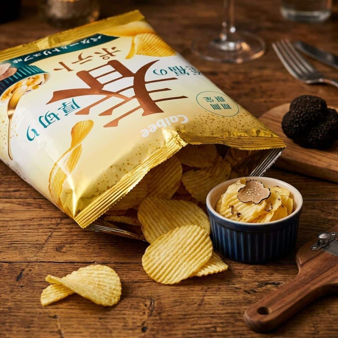Calbee Thick Cut Butter Truffle Potato Chips 1 | Snack Affair Archives Bag of ridged potato chips spilled on a wooden table with chips scattered and a small cup of creamy dip topped with flakes nearby.