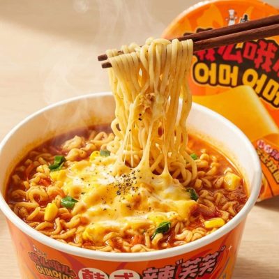 Chopsticks lift steaming noodles from an orange-cup ramen with melted cheese, corn, and green onions.