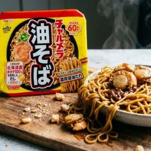 Archives Yellow Japanese instant noodle box beside a plate of saucy noodles with scallops on a wooden board, steam rising in the background.