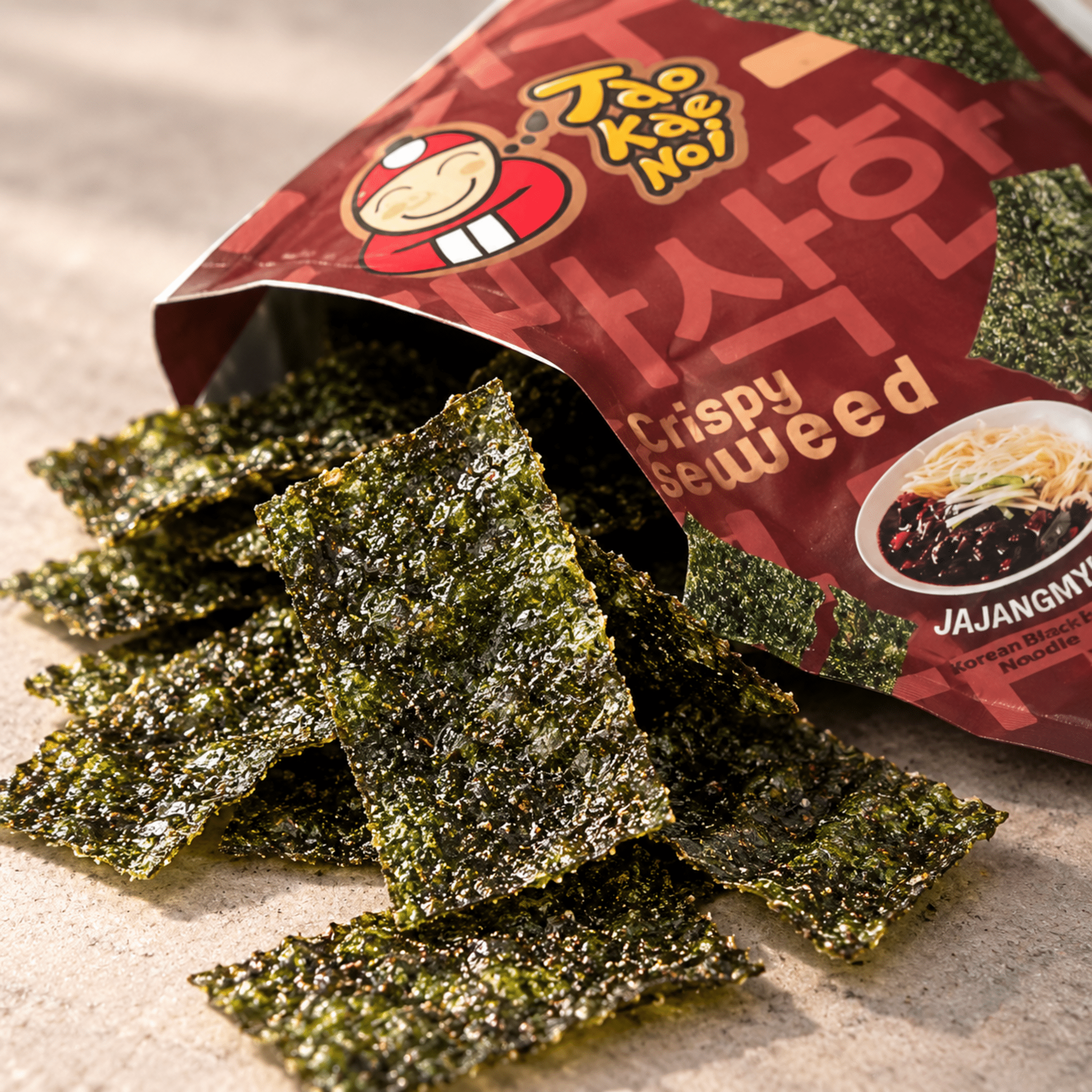 Tao Kae Noi Jajangmyeon Crispy Seaweed 32g Close Up | Snack Affair Archives Crispy seaweed sheets scattered on a light surface next to a red snack bag with a cartoon logo.
