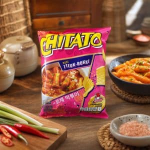 Bright pink bag of Chitato snacks on a wooden table, surrounded by fresh chili peppers, lemongrass, and a bowl of seasoned noodles.