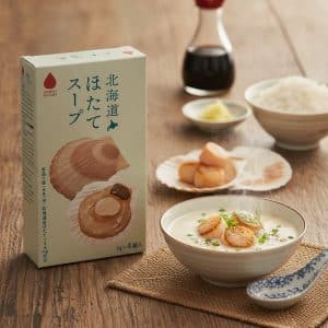 Box of Hokkaido scallop soup mix beside a bowl of creamy scallop soup and a plate of scallops on a wooden table.