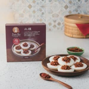 Boxed Jian Bo dim sum beside a plate of white steamed rice cakes topped with spicy minced meat on a kitchen counter.