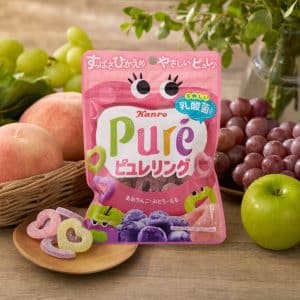 Pink Kanro Pure fruit gummies bag on a wooden table, surrounded by peaches, grapes, and a green apple.
