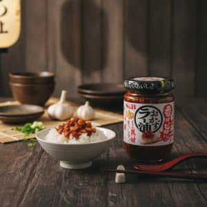 Jar of chili oil beside a bowl of white rice topped with crispy bits; garlic and bowls in the background.