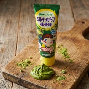 Archives Milky matcha candy tube standing upright on a wooden board with a dollop of green matcha paste in the foreground and powder scattered around.