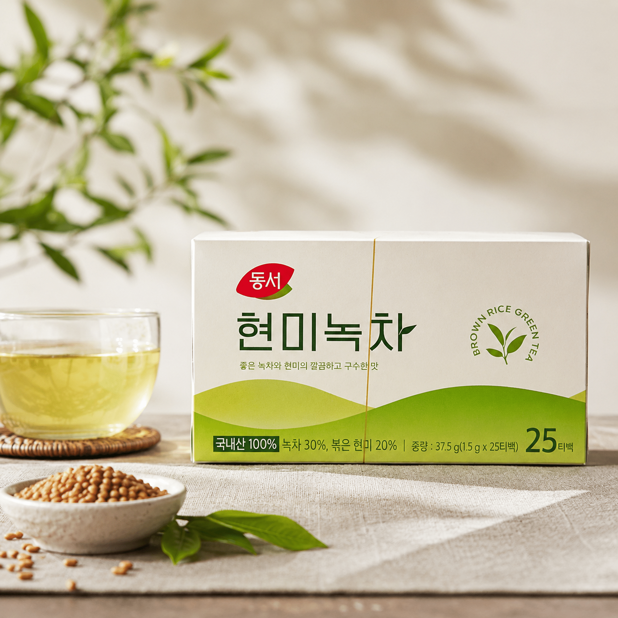 DongsuhBrownRiceGreenTea25TeaBagsLifestylepng | Snack Affair Archives Front-right close-up of a white box with green design and Korean text on a light table, with a small bowl of crunchy grains in the foreground and a blurred leafy plant in the background.