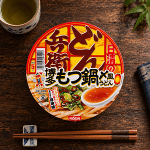 Archives Top-down view of a Nissin instant ramen cup with red and yellow packaging, on a dark wooden table, chopsticks and a small dish nearby.
