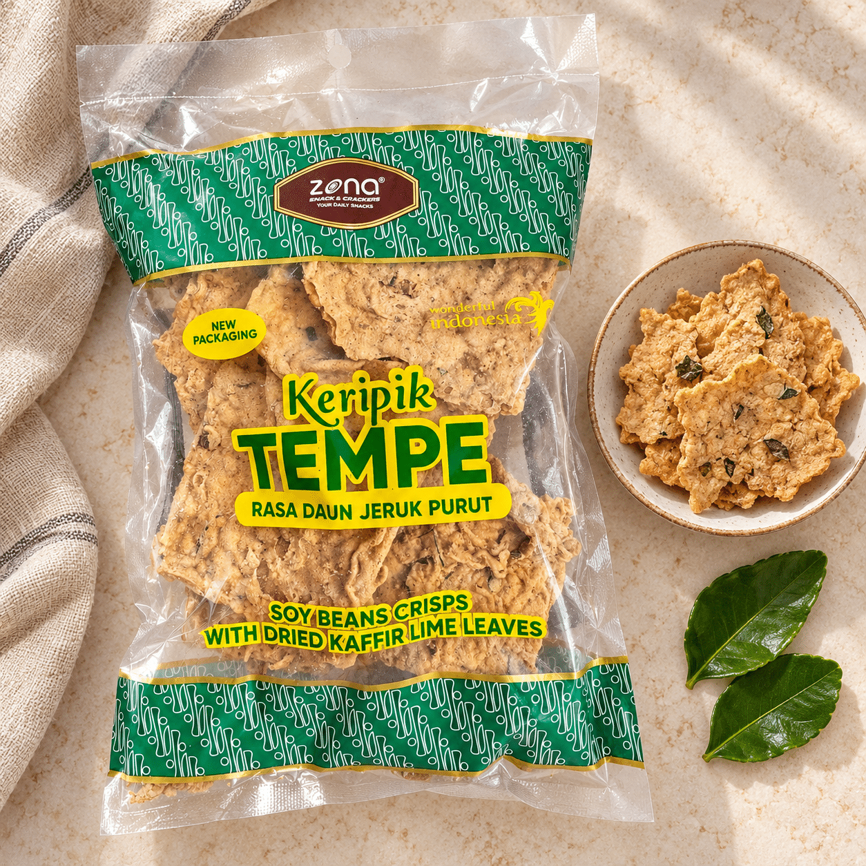 ZONA kaffir lime tempe crisps | Snack Affair Archives Front of a green and yellow bag of Keripik Tempe soy bean crisps with dried kaffir lime leaves, on a beige countertop with a bowl of chips nearby.