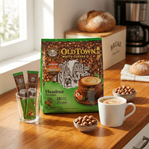Box of OldTown White Coffee Hazelnut flavor on a kitchen counter with a white mug of coffee and bowls of nuts nearby, bathed in natural light